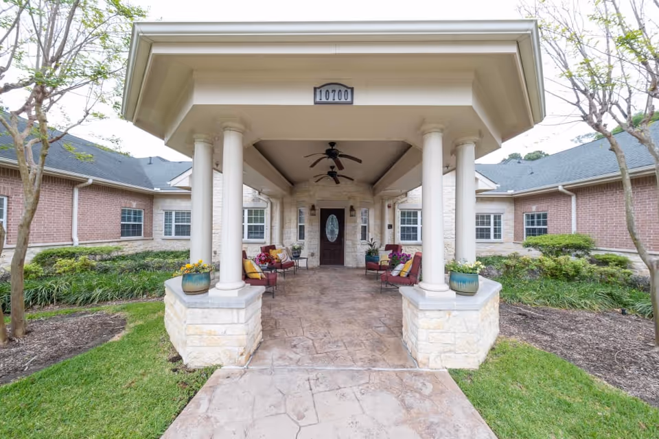Covered front entrance of a brick assisted living building with columns, outdoor seating, potted plants and an address plaque reading "10700".
