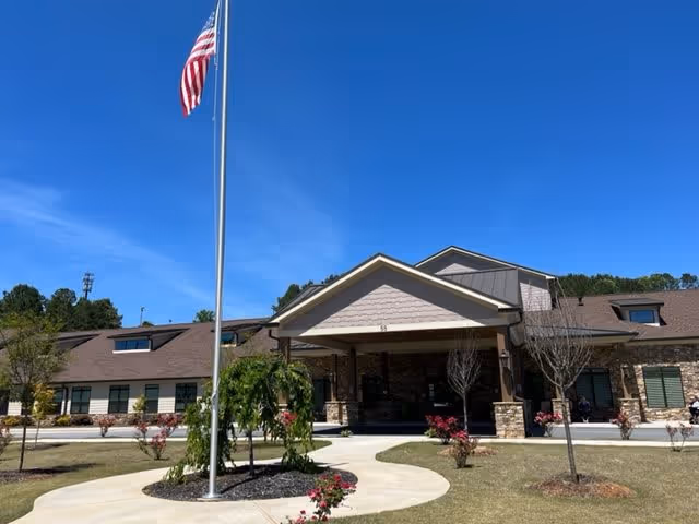 Exterior view of Dahlonega Assisted Living & Memory Care facility showing a single-story building with a covered entrance, stone and siding facade, landscaped grounds with small trees and bushes, a curved walkway, and an American flag on a flagpole against a clear blue sky.