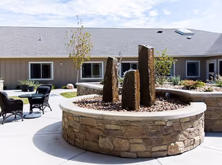 Outdoor courtyard with round stone planters featuring vertical stone water features, patio tables and chairs, and a single-story building in the background.
