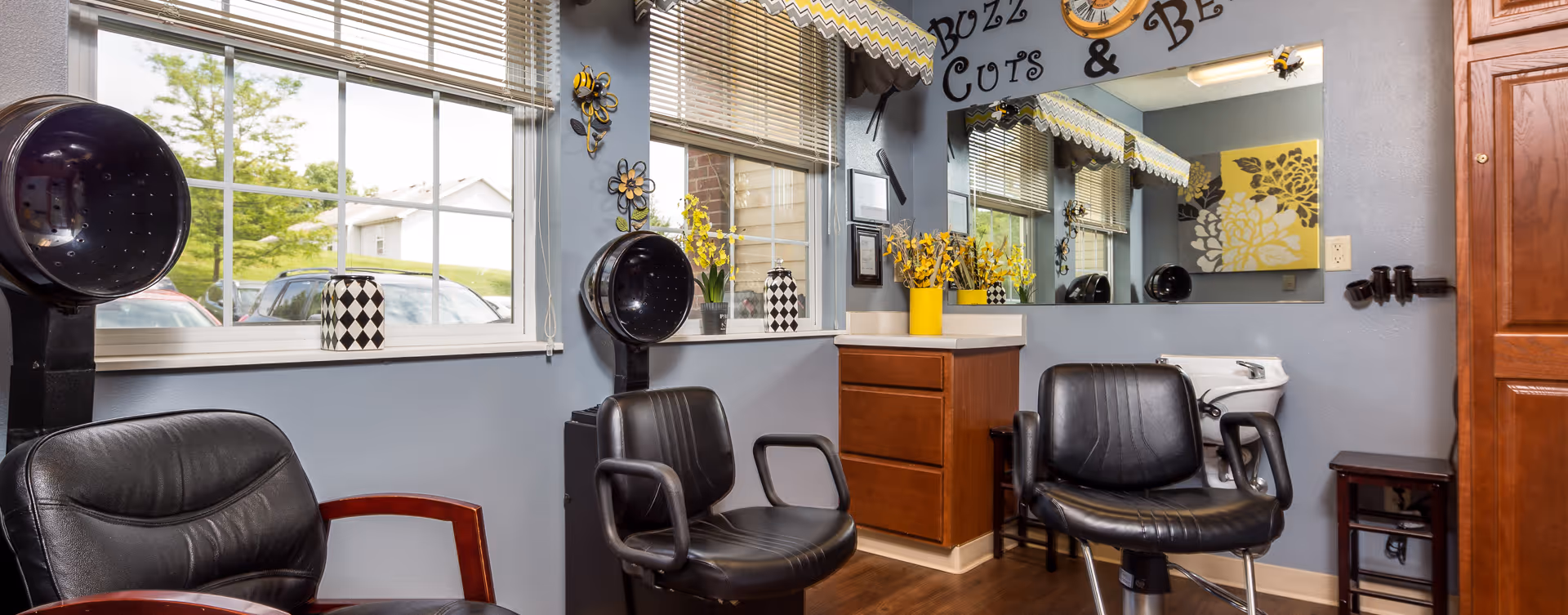 Interior of a hair salon area in a senior living facility with two black salon chairs, hair dryers, a large mirror, and decorative yellow flowers. The walls are painted gray with windows showing a view of parked cars and greenery outside. The words 'Buzz Cuts & Be' are partially visible above the mirror.