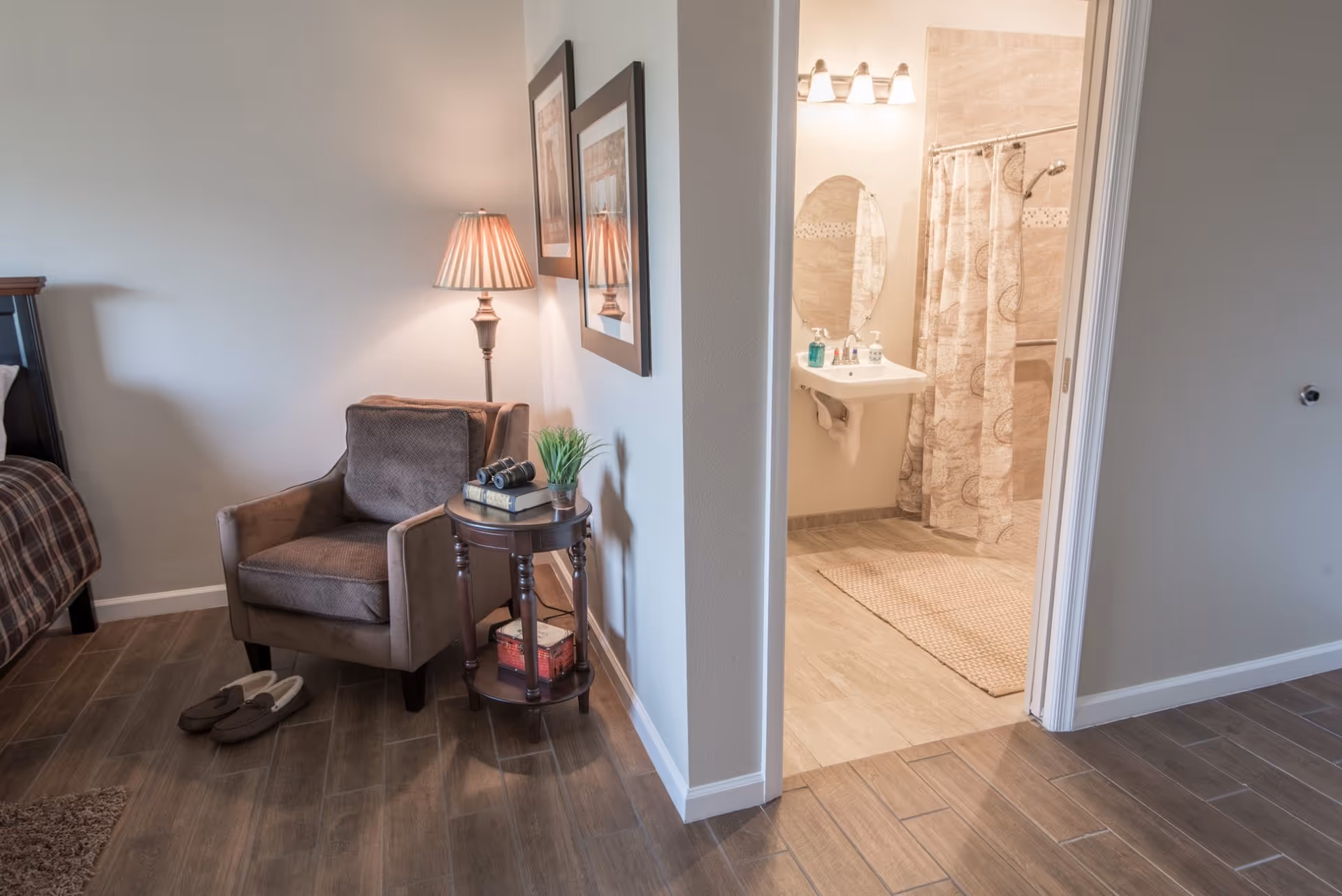 A cozy corner of a senior living facility room with a brown armchair, a small round wooden side table holding books, a plant, and binoculars, a floor lamp with a pleated shade, and two framed pictures on the wall. To the right, an open doorway reveals a bathroom with a pedestal sink, an oval mirror, a soap dispenser, and a shower with a patterned curtain.