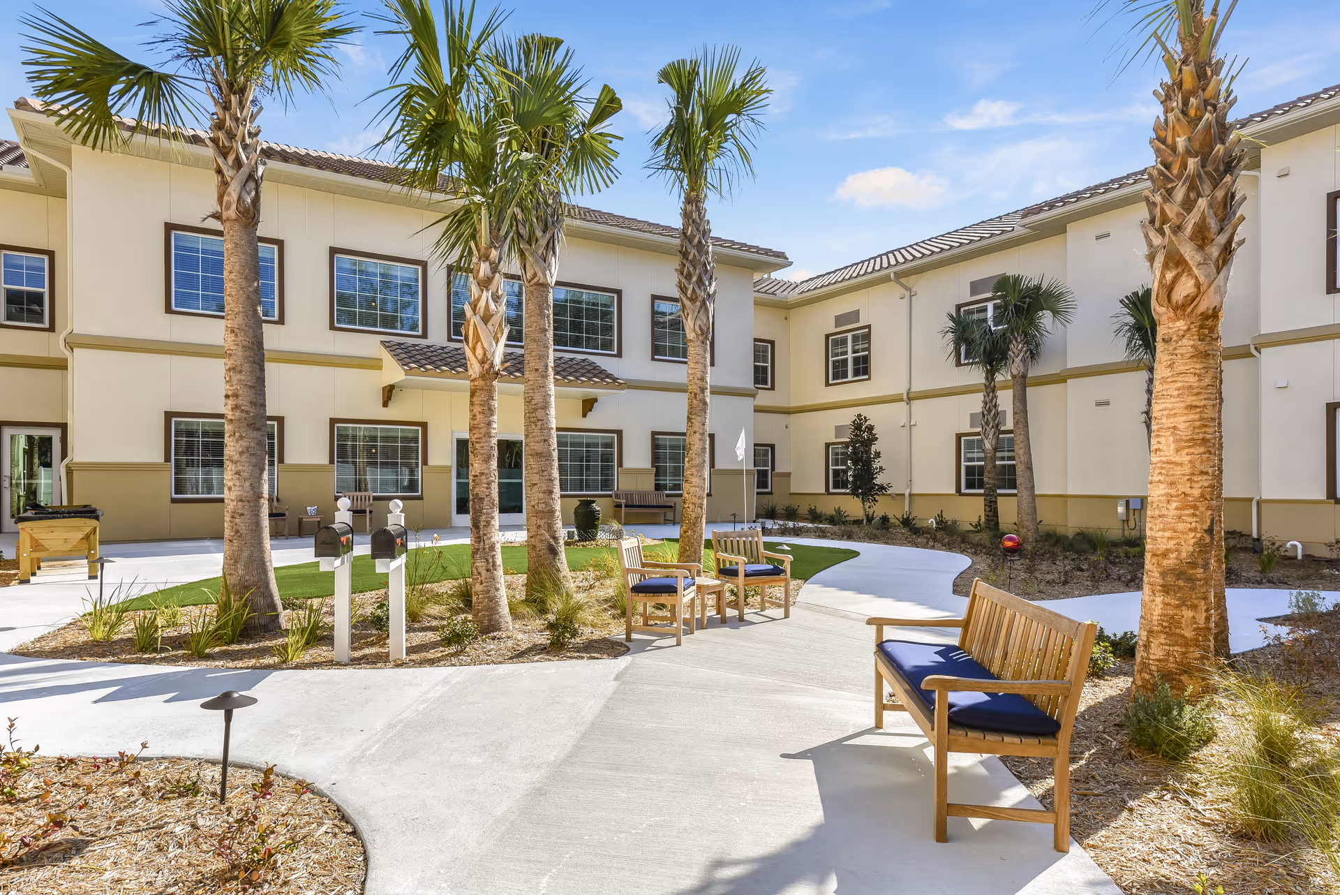 Outdoor courtyard area at Tuscan Gardens of Palm Coast Senior Living featuring palm trees, wooden benches with blue cushions, a curved concrete walkway, and a two-story beige building with multiple windows in the background under a blue sky with some clouds.