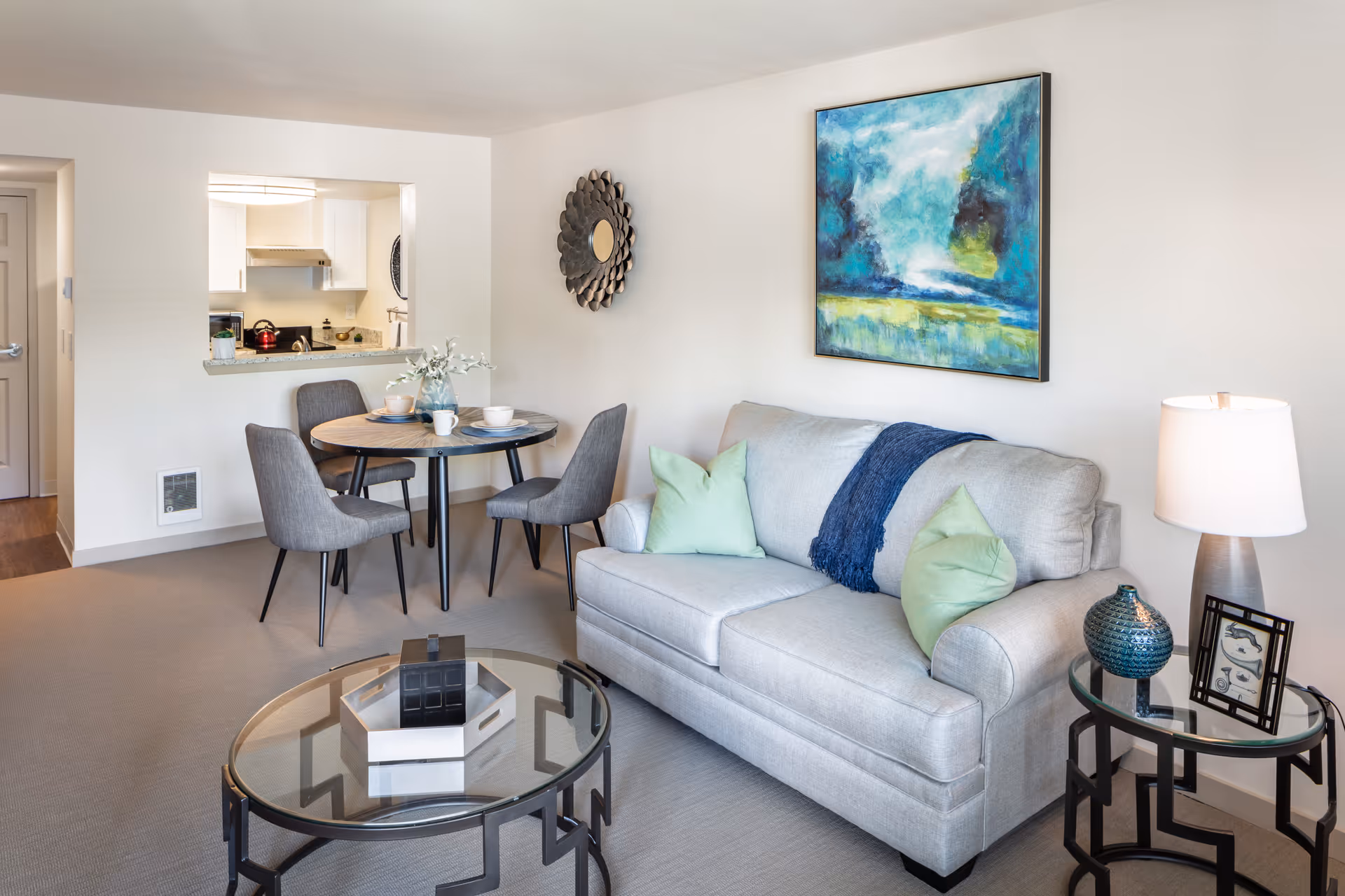 Bright furnished living room with a gray sofa, glass coffee table, side table with a lamp, and a small dining table with chairs beside a pass-through to the kitchen.