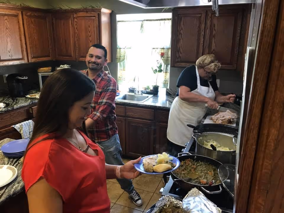 A woman in a red shirt holding a blue plate with food in a kitchen. A man in a red plaid shirt stands behind her smiling. Another woman wearing an apron is carving meat at the counter. The kitchen has wooden cabinets, a granite countertop, and pots of food on the stove.