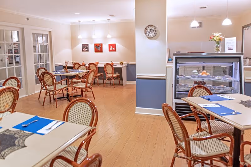 A cozy dining area in a senior living facility with several tables and chairs arranged neatly. The tables are set with blue placemats, silverware, and napkins. The room features light-colored walls with a blue wainscoting, wooden flooring, and soft overhead lighting. There is a display case with pastries and a vase with flowers on top. Three small framed pictures hang on the far wall, and a clock is mounted on a column in the center of the room.