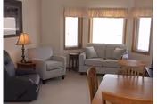 A cozy living room with a gray loveseat and matching armchair, a black recliner, a wooden side table with a lamp, and a wooden dining table with chairs. Three windows with beige curtains allow natural light into the room.