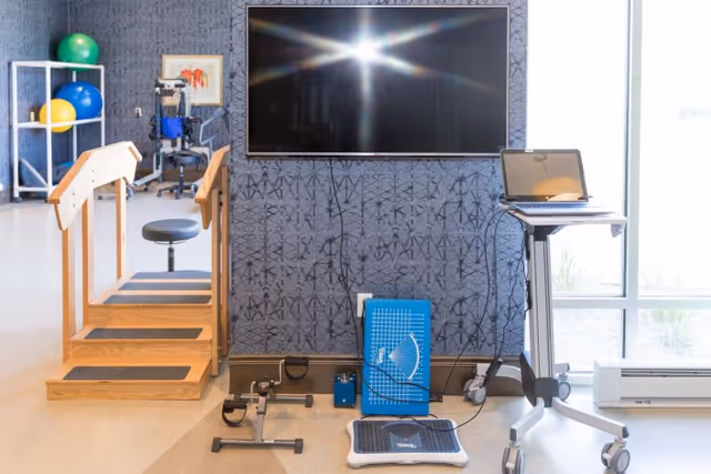Physical therapy room with wooden stairs for rehabilitation, exercise equipment including a pedal exerciser, balance board, therapy balls on a white rack, a large flat-screen TV mounted on a patterned wall, and a laptop on a mobile stand near a window.