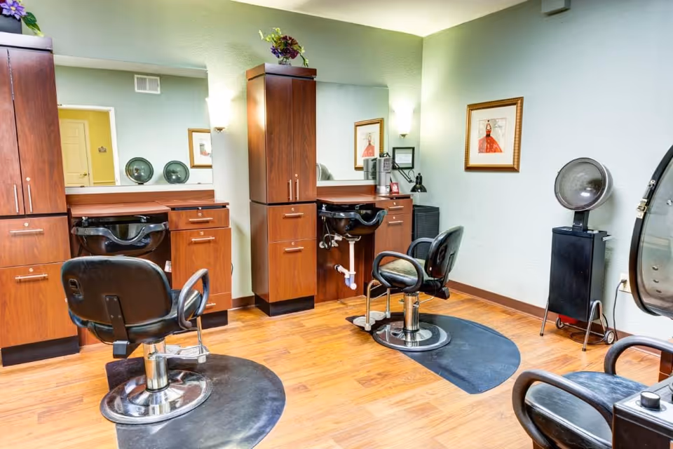 Interior of a hair salon area with two black salon chairs in front of wooden cabinets and sinks for washing hair. The room has light green walls, wooden flooring, a large mirror, framed artwork, and a hair dryer on a stand.