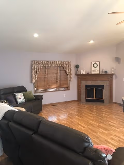 A living room with light purple walls and wooden flooring. There are two black leather sofas with cushions, a window with wooden blinds and floral valance, and a wooden fireplace mantel decorated with framed art, a small plant, and decorative items.
