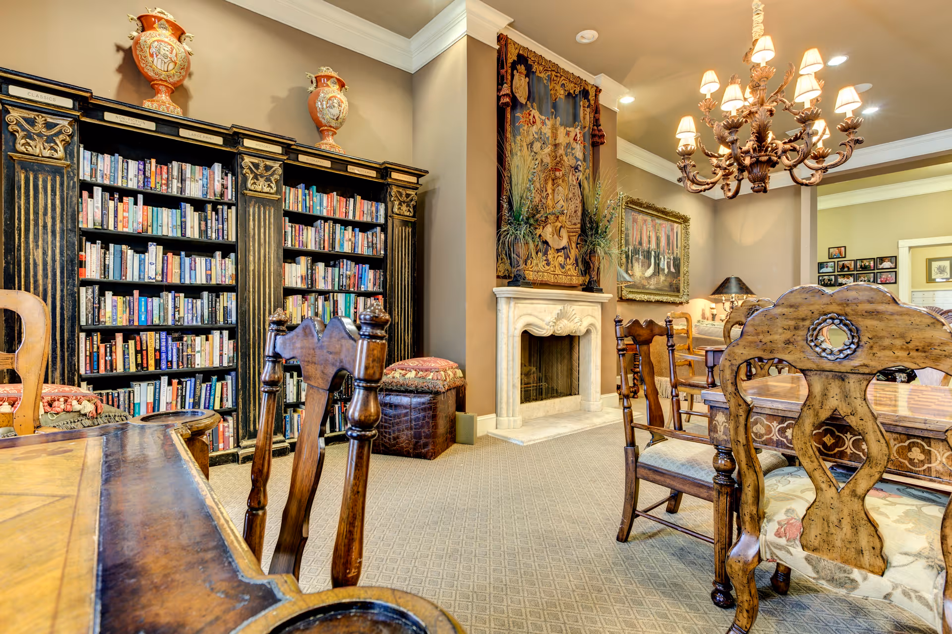 A cozy interior room featuring a large ornate bookshelf filled with books, antique wooden chairs and tables, a decorative fireplace with a tapestry above it, and a chandelier hanging from the ceiling. The room has warm lighting and classic decor.