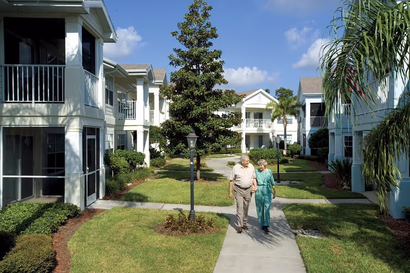 An elderly couple walking hand in hand on a paved pathway between two rows of white residential buildings with balconies, surrounded by green lawns, trees, and shrubs under a blue sky with some clouds.