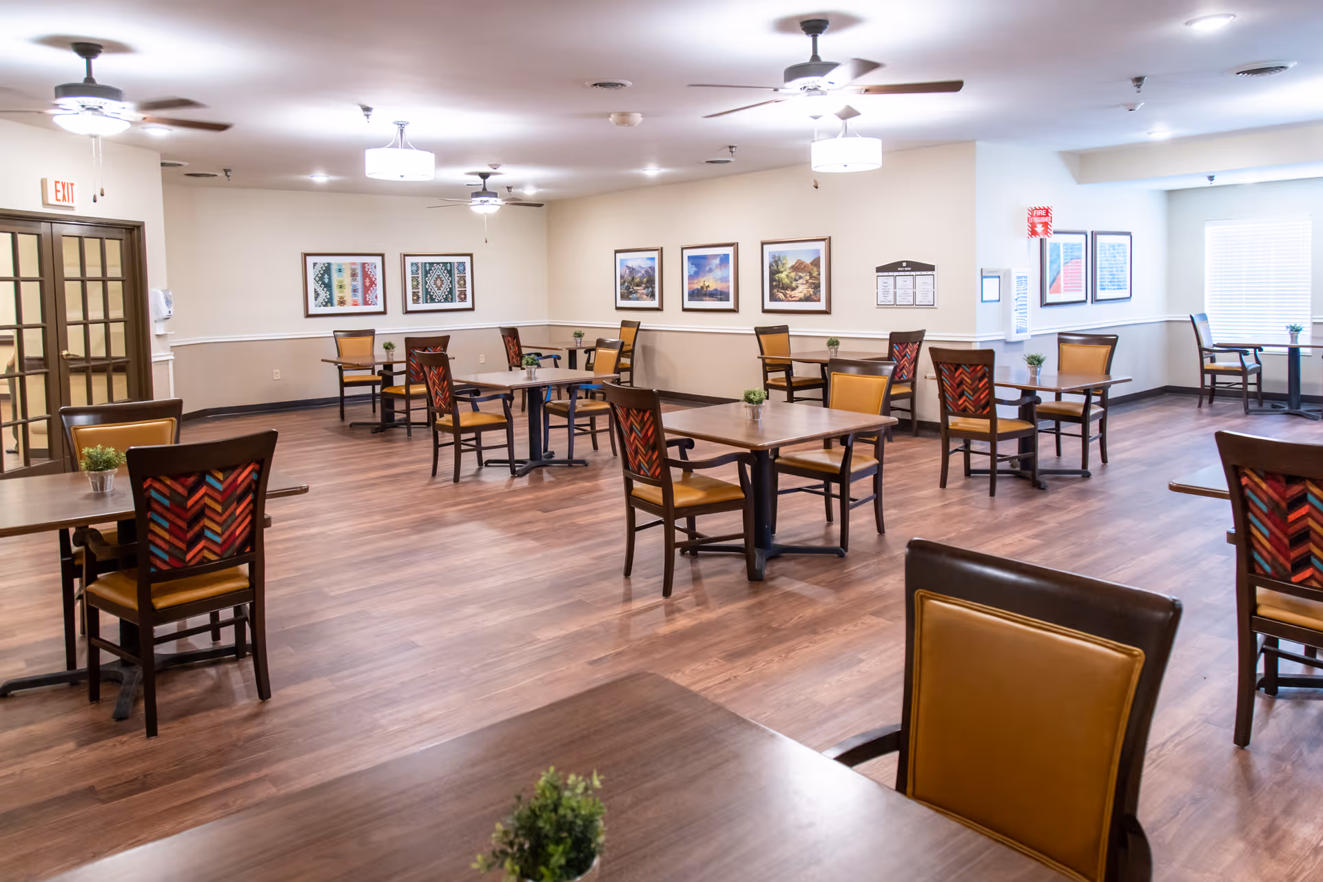 A spacious dining room with wooden floors and multiple tables and chairs arranged neatly. The chairs have colorful patterned backs and tan seats. The walls are decorated with framed artwork, and there are ceiling fans and light fixtures overhead. Small potted plants are placed on the tables. Large windows allow natural light to brighten the room.
