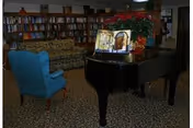 A cozy interior room featuring a black grand piano with an open music book on its stand, a blue upholstered armchair, a patterned sofa, and a large bookshelf filled with books along the back wall. There is also a large potted plant with red flowers on the piano.