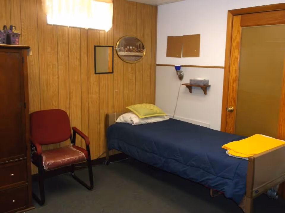 Small bedroom with a single bed covered in a dark blue comforter, a red chair, wood-paneled walls, and a wooden door.