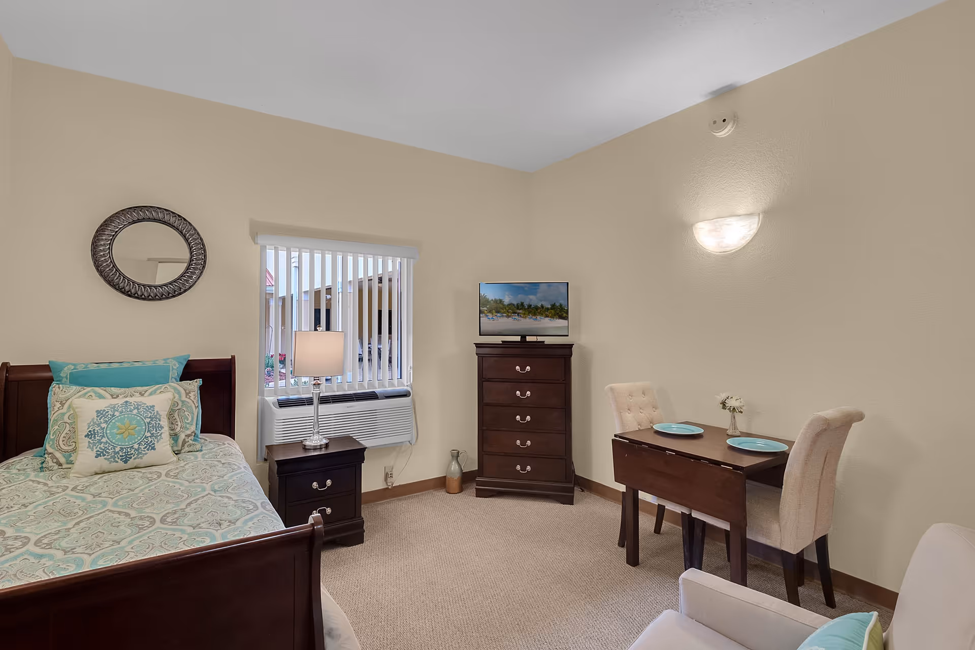A cozy assisted living room featuring a single bed with patterned bedding and multiple pillows, a nightstand with a lamp, a window with vertical blinds, a tall dresser with a TV on top, a small dining table set with two chairs and two plates, and a comfortable armchair in the foreground.
