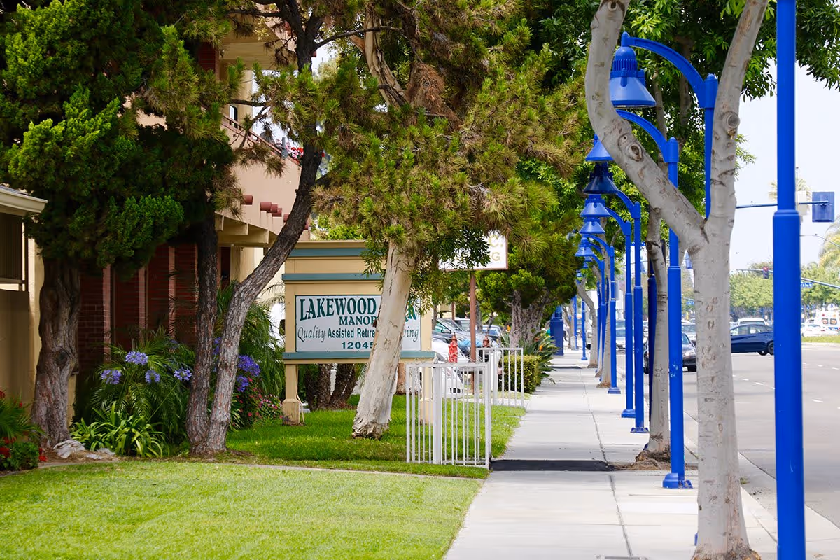 Sidewalk lined with trees and blue street lamps running alongside a street. On the left side, there is a green lawn and a sign that reads 'Lakewood Manor Quality Assisted Retirement Living'.