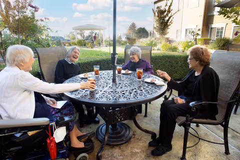 Four elderly women seated around a metal patio table outdoors, laughing and enjoying iced tea and snacks.