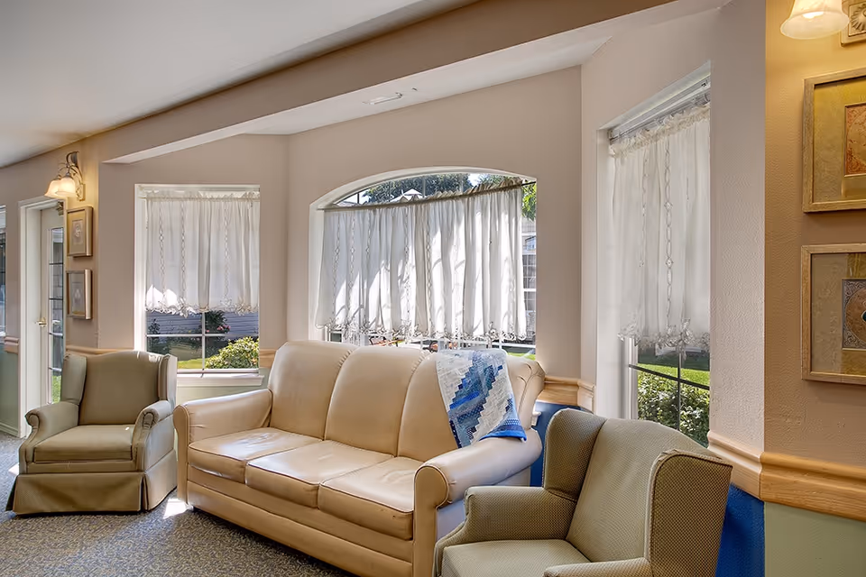 A cozy living room area with a beige leather sofa and two green upholstered armchairs. The room has large windows with white lace curtains allowing natural light to brighten the space. There are framed pictures on the wall and a quilt draped over the sofa's armrest.
