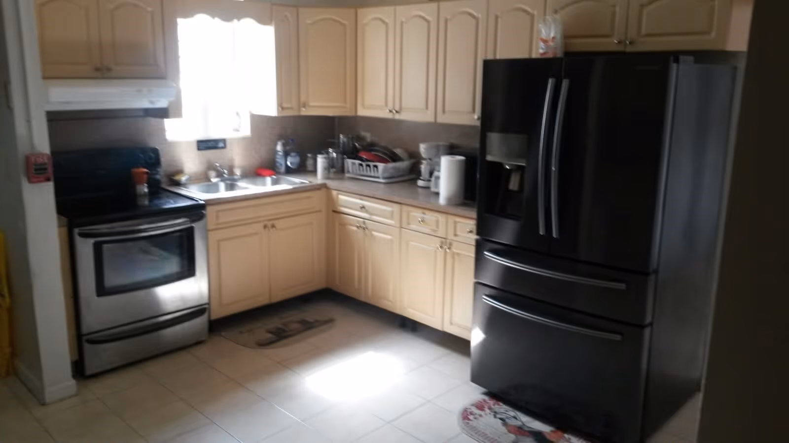 A kitchen with cream-colored cabinets, a stainless steel oven and stove on the left, a double sink under a window, and a large black refrigerator with French doors on the right. Various kitchen items such as a dish rack, paper towel roll, and coffee maker are on the countertops.