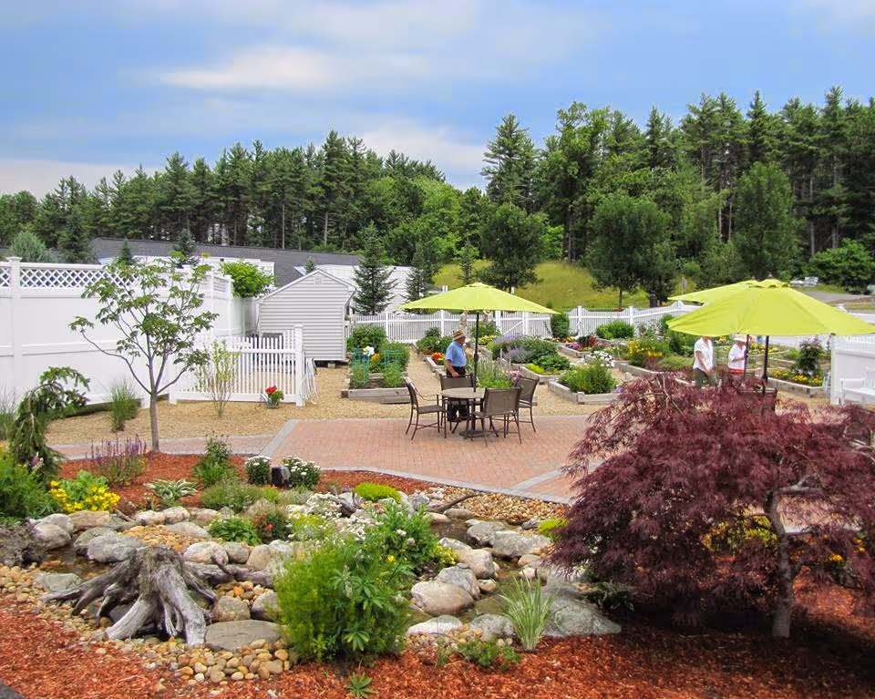 A landscaped courtyard with patio tables and lime-green umbrellas surrounded by raised garden beds, trees, and rock features.