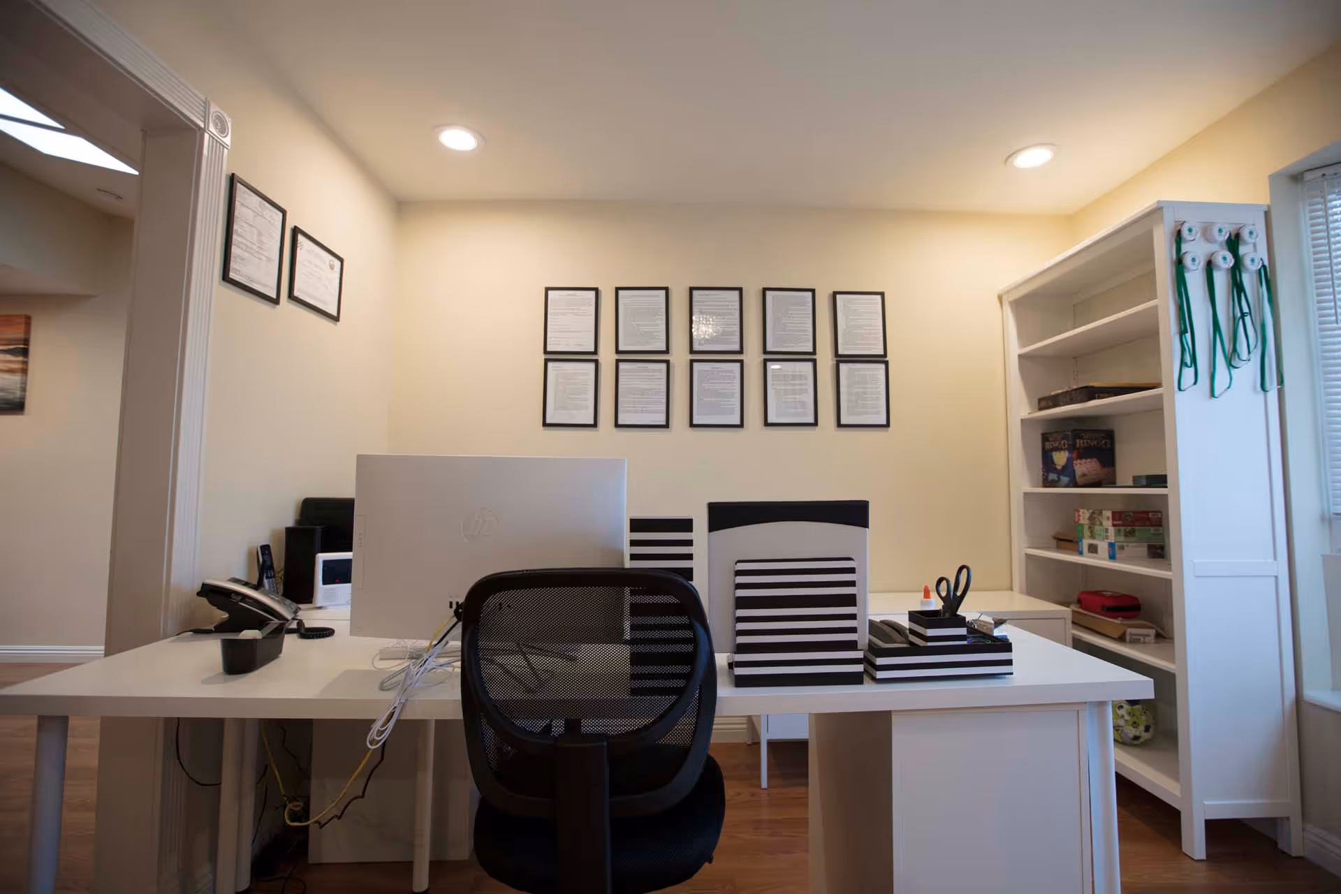 Bright office/reception area with a desk, computer monitor, office chair, framed documents on the wall, and shelving with supplies.