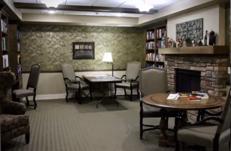 A cozy senior living common area with a stone fireplace on the right, decorated with small sculptures and a metal wall art piece above the mantle. There are two round wooden tables surrounded by cushioned chairs, a floor lamp, and bookshelves filled with books. The walls have a textured green wallpaper and the carpet is light gray.
