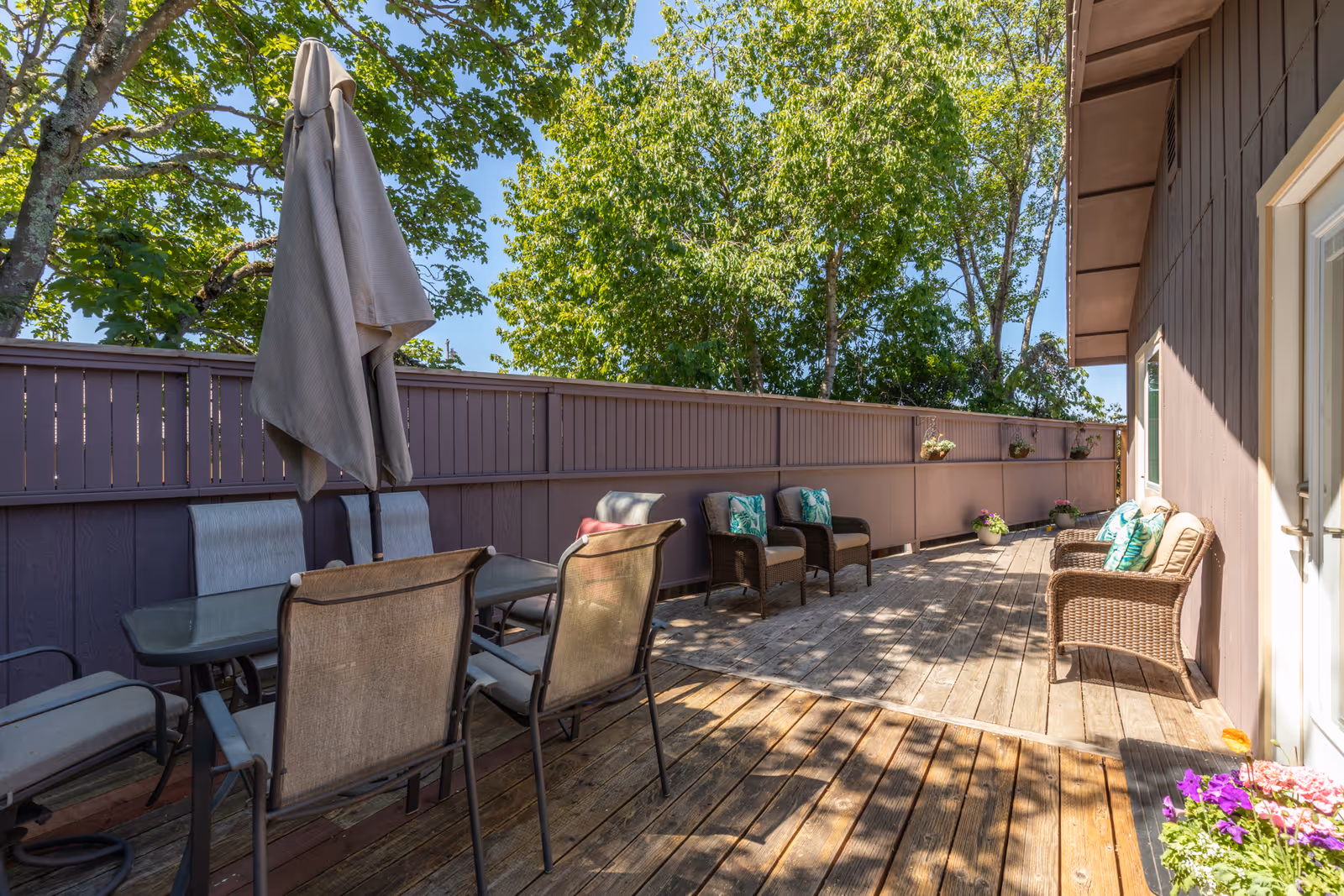 Wooden outdoor deck with a dining table and chairs, wicker seating, potted flowers, and a tall privacy fence under trees.