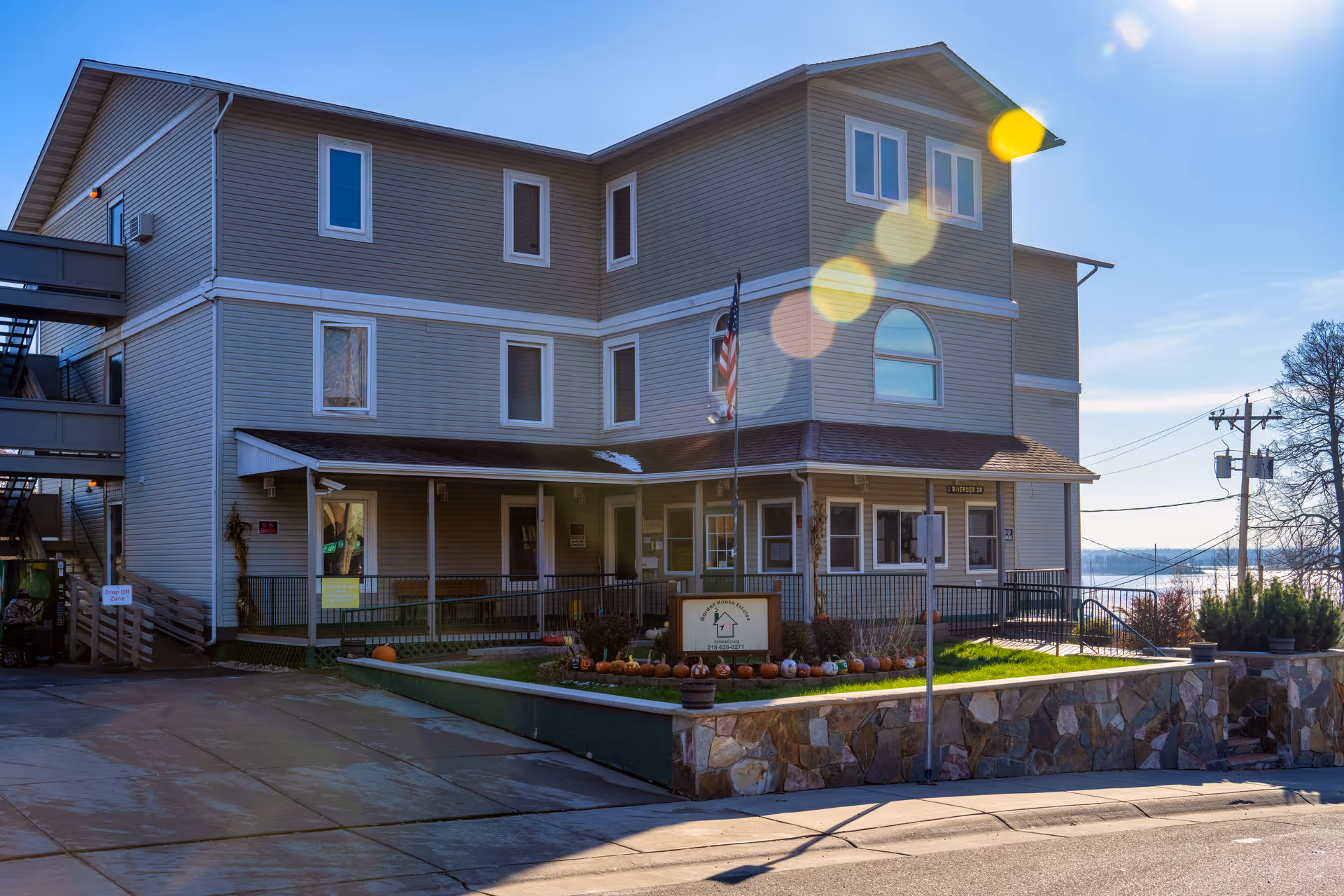 Exterior view of a three-story senior living facility building with beige siding, multiple windows, and a covered entrance. There is a small garden area with pumpkins and a sign in front of the building. An American flag is displayed on a flagpole near the entrance. The sky is clear with sunlight creating lens flare effects in the image.
