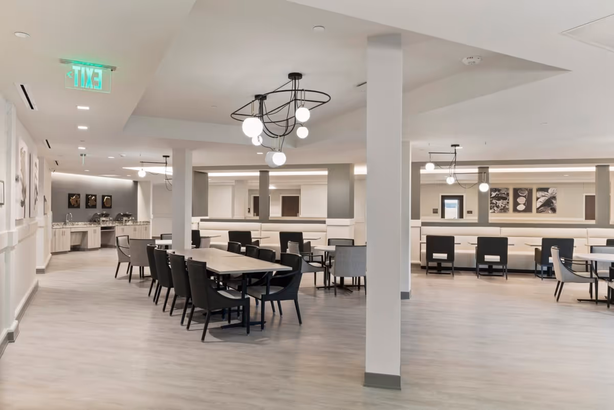 A spacious and modern dining area with multiple tables and chairs arranged neatly. The room features light-colored flooring, white walls, and contemporary light fixtures hanging from the ceiling. There are artworks on the walls and a counter area with sinks and serving stations in the background.