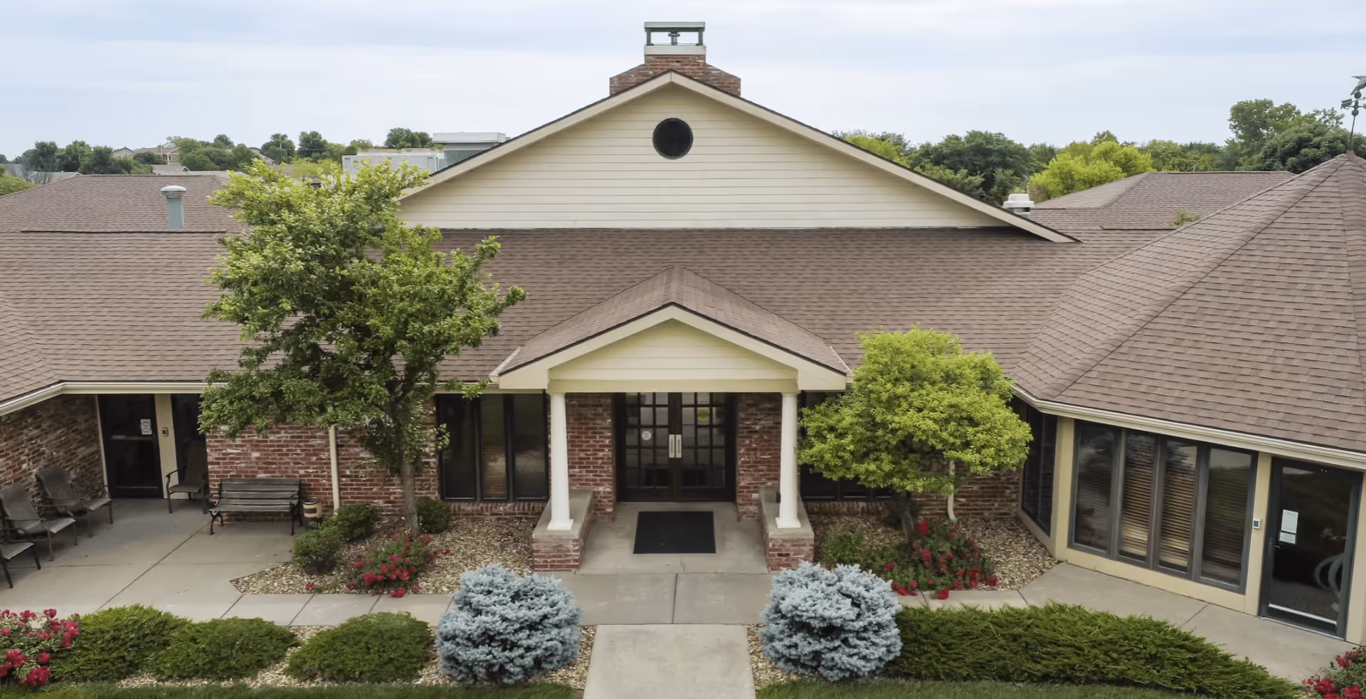 Front entrance of a single-story brick assisted living building with a covered portico, flanked by trees and landscaping.