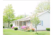 Single-story light-colored cottage with a front porch, lawn, flowering shrubs, and trees.