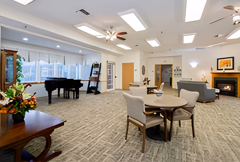 A spacious and well-lit common area in a senior living facility featuring a round table with four chairs, a grand piano near large windows, a cozy seating area with sofas around a fireplace, ceiling fans, and decorative plants and artwork.