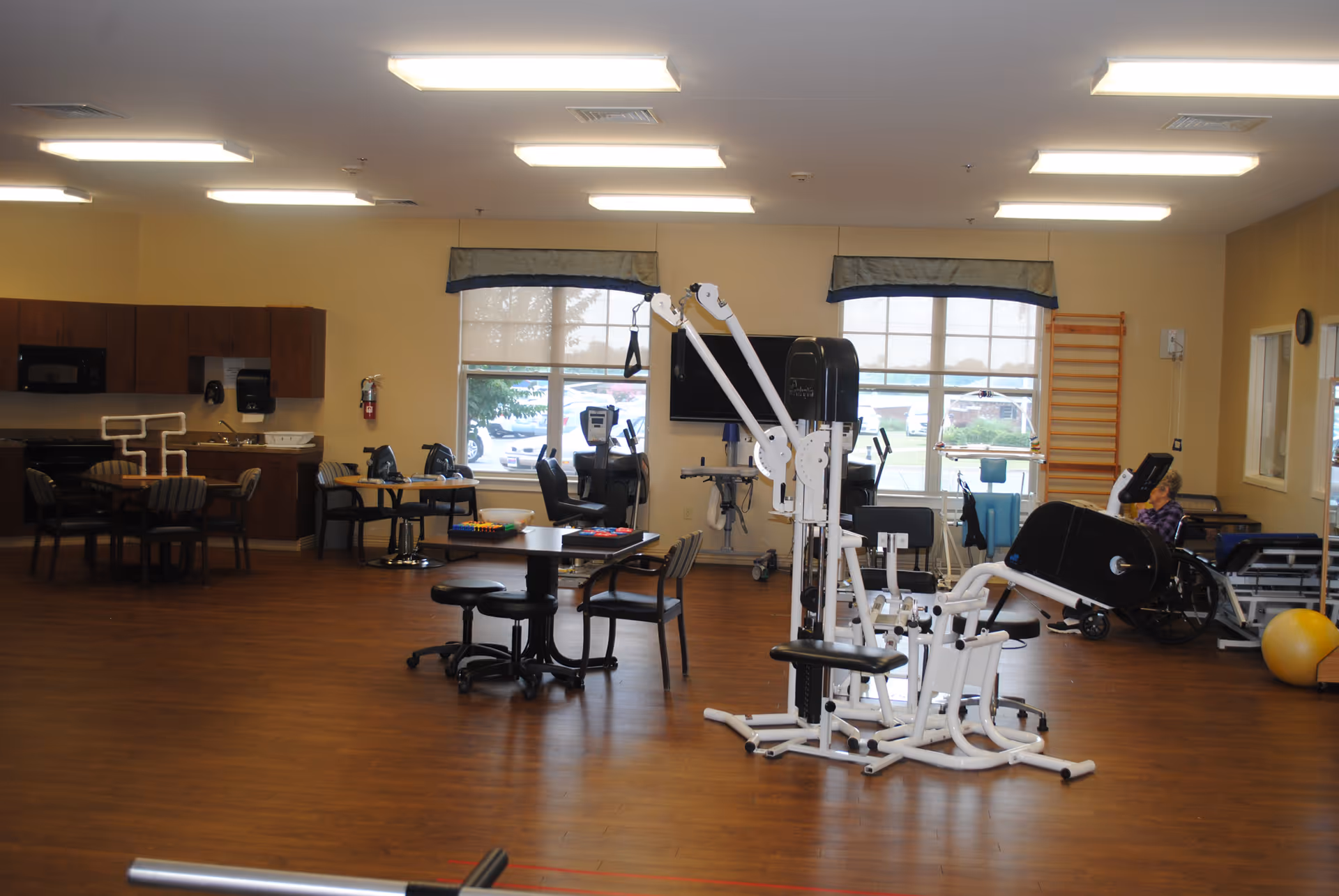 A spacious room with wooden flooring featuring exercise equipment, tables with chairs, and a kitchenette area with cabinets, a microwave, and a sink. Large windows with valances allow natural light to enter. A person in a wheelchair is visible near the back of the room.