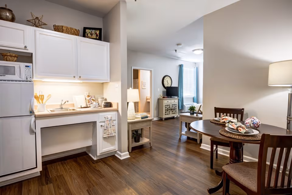 Interior view of a senior living facility apartment showing a small kitchen area with white cabinets, a refrigerator, microwave, and sink. Adjacent to the kitchen is a dining area with a round wooden table set for two, and beyond that is a living room with a TV, clock on the wall, and a window with blue curtains. The flooring is wood and the walls are painted light gray.