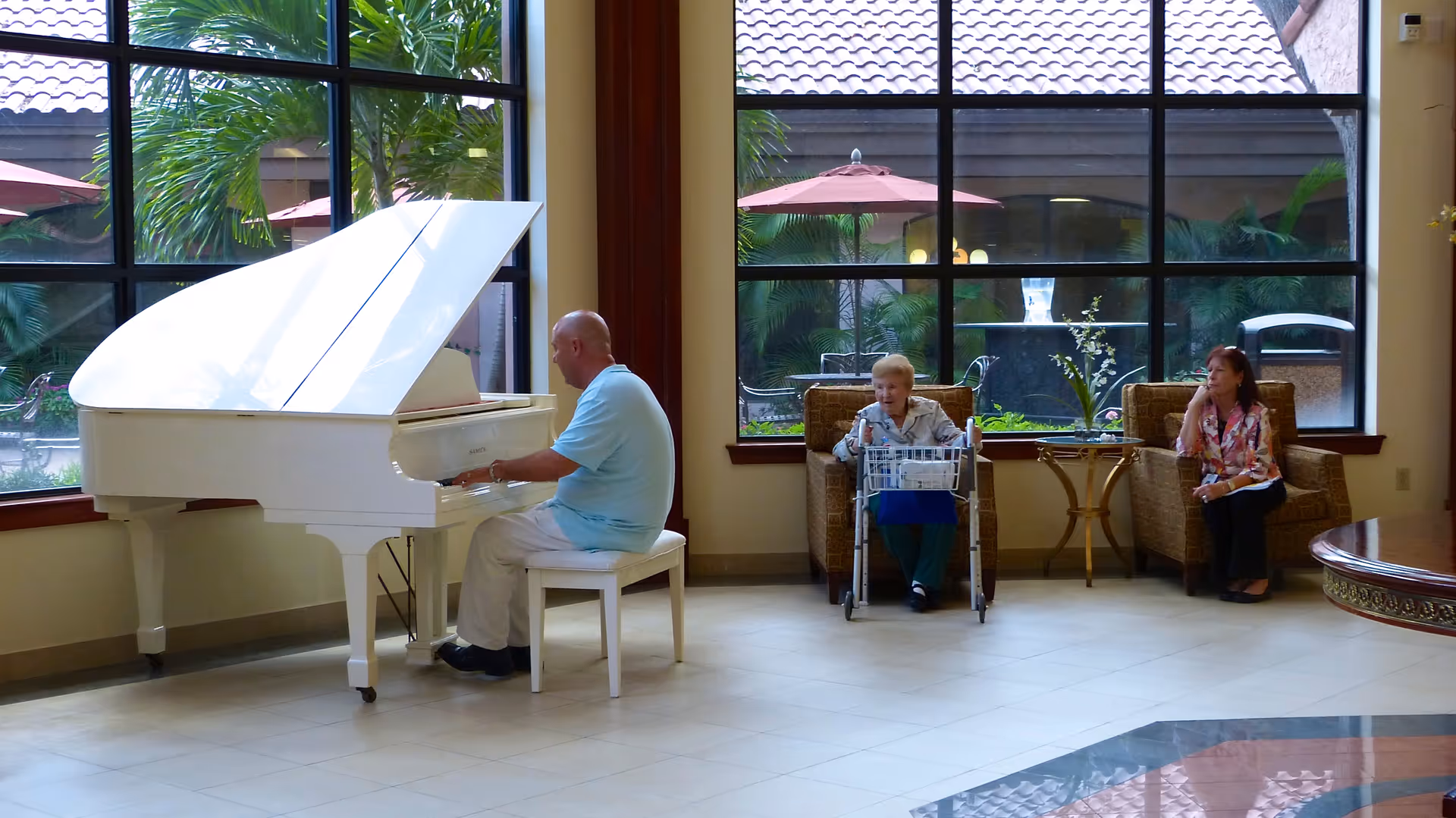A man in a light blue shirt is playing a white grand piano in a spacious room with large windows. Two women are seated on armchairs nearby, one with a walker in front of her and the other sitting with her hand on her face. Outside the windows, there are palm trees and patio umbrellas.