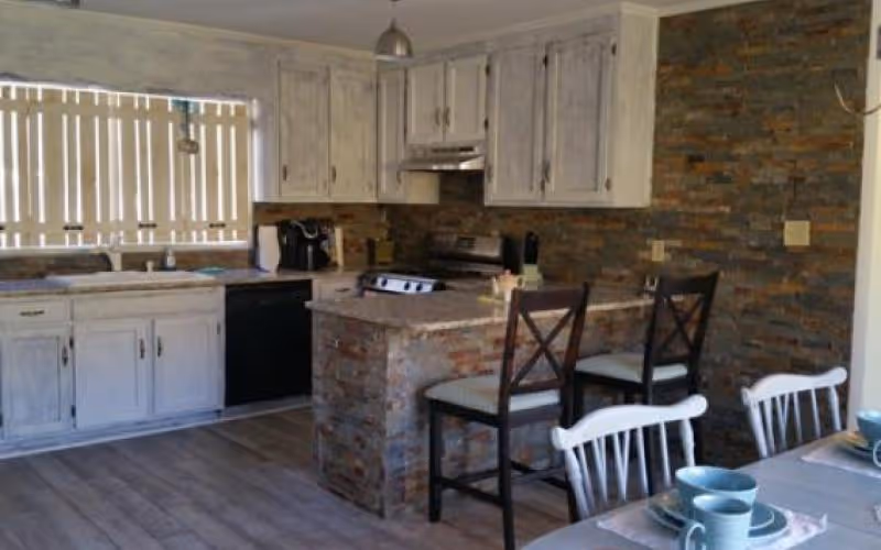 A kitchen with white distressed cabinets, a stone brick island with two chairs, a stove, dishwasher, and a dining table set with blue cups and plates. The walls have a stone brick finish and there is a window with vertical blinds.