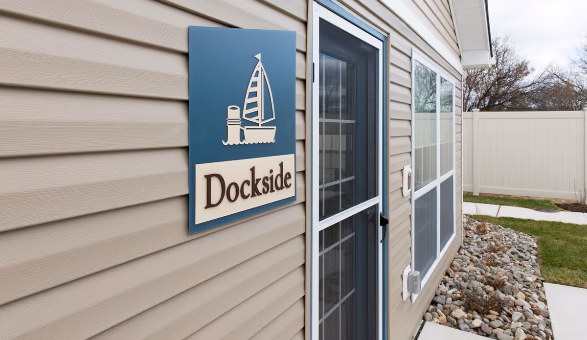 Exterior view of a beige siding building with a blue and white sign that reads 'Dockside' featuring a sailboat and lighthouse graphic. There is a glass door with a black handle and two large windows next to it. The ground is covered with rocks and a concrete pathway, with a white fence and some trees in the background.