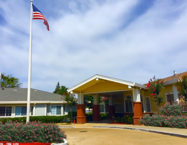 Entrance of Gramercy Court Skilled Nursing & Rehabilitation Center with a covered driveway supported by brick pillars, surrounded by bushes and flowering plants, and an American flag on a flagpole against a partly cloudy sky.