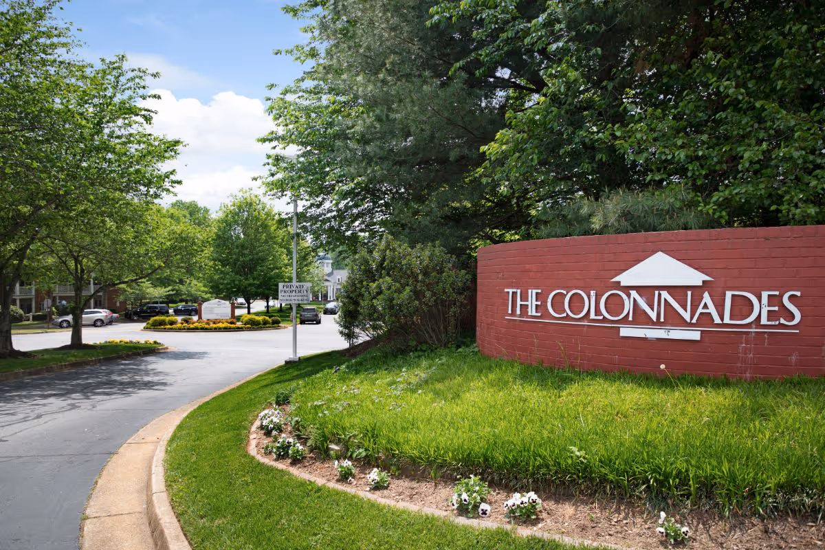 Brick entrance sign reading 'THE COLONNADES' beside a landscaped driveway and trees.