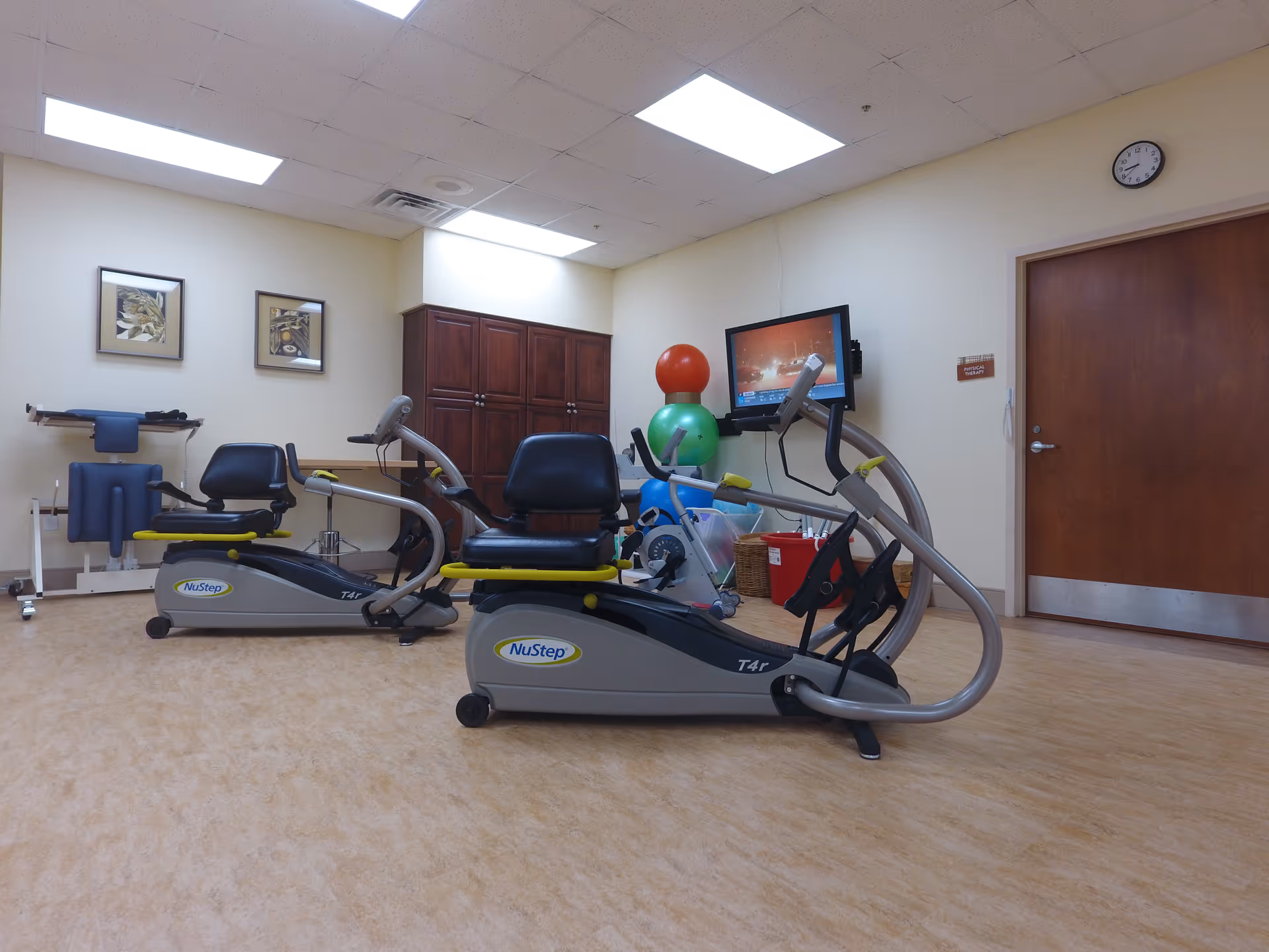 A room with two NuStep T4r recumbent cross trainers positioned on a light brown floor. The room has beige walls with two framed pictures, a wooden cabinet, and a wall-mounted TV displaying a program. There are colorful exercise balls stacked in the corner and a clock above a closed wooden door.