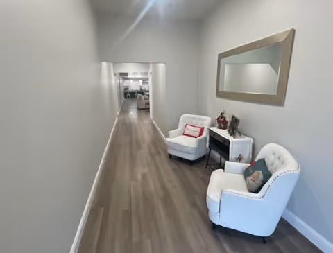 Long narrow hallway with two white armchairs, a small side table and mirror along the right wall leading toward a seating area.
