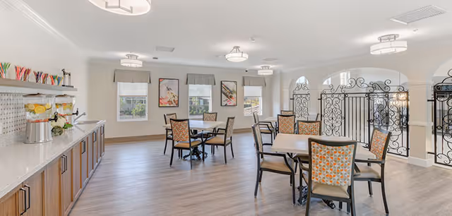 Bright communal dining room with several tables and chairs, a beverage counter, and decorative wrought-iron railings.