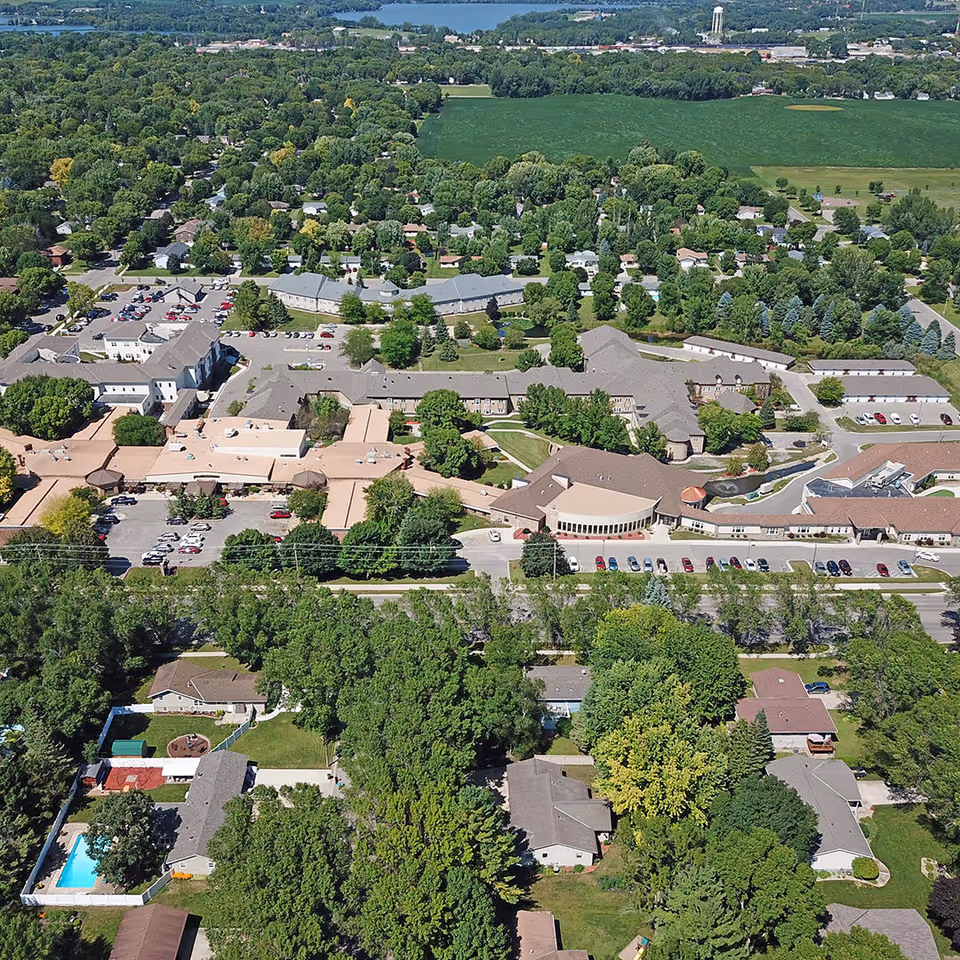 Aerial view of a senior living facility named Bethesda surrounded by trees and residential houses, with parking lots and green fields in the background.