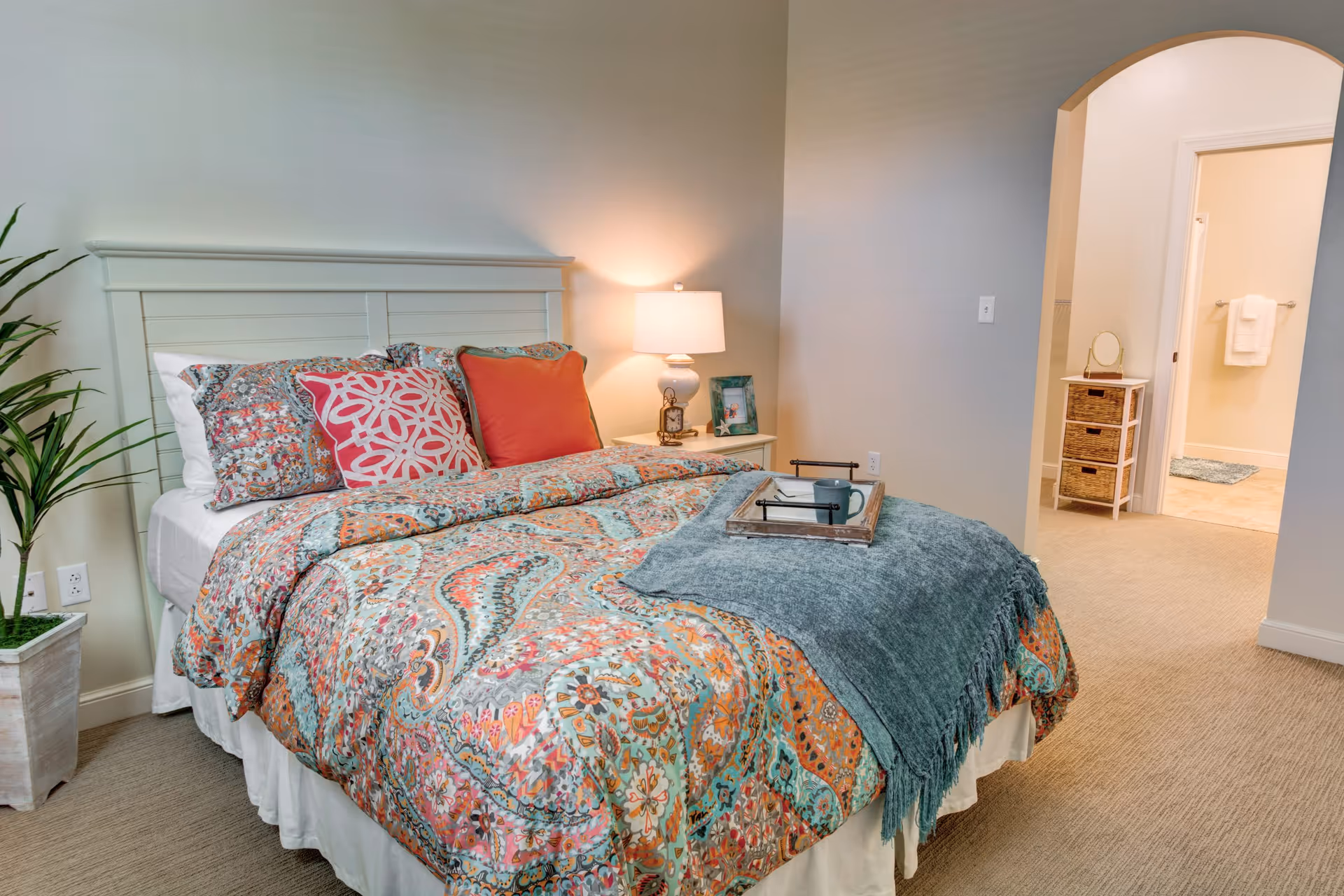 Well-lit bedroom with a patterned comforter on a bed, a bedside table and lamp, a potted plant, and an arched doorway leading to an adjoining bathroom area.