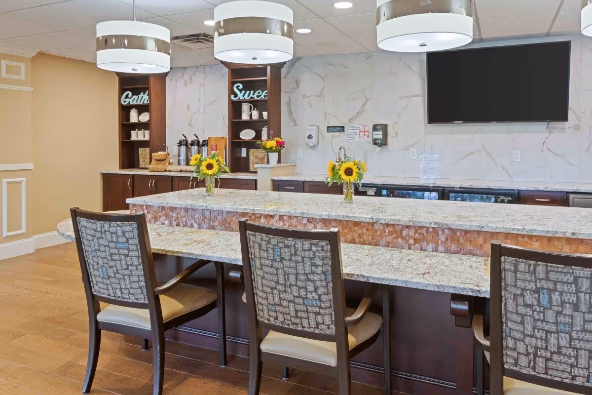 Interior view of a modern kitchen or dining area with a long granite countertop island, three chairs with patterned backs, two vases with sunflowers on the counter, wooden cabinets, a wall-mounted flat screen TV, and decorative shelves with the words 'Gather' and 'Sweet' displayed.