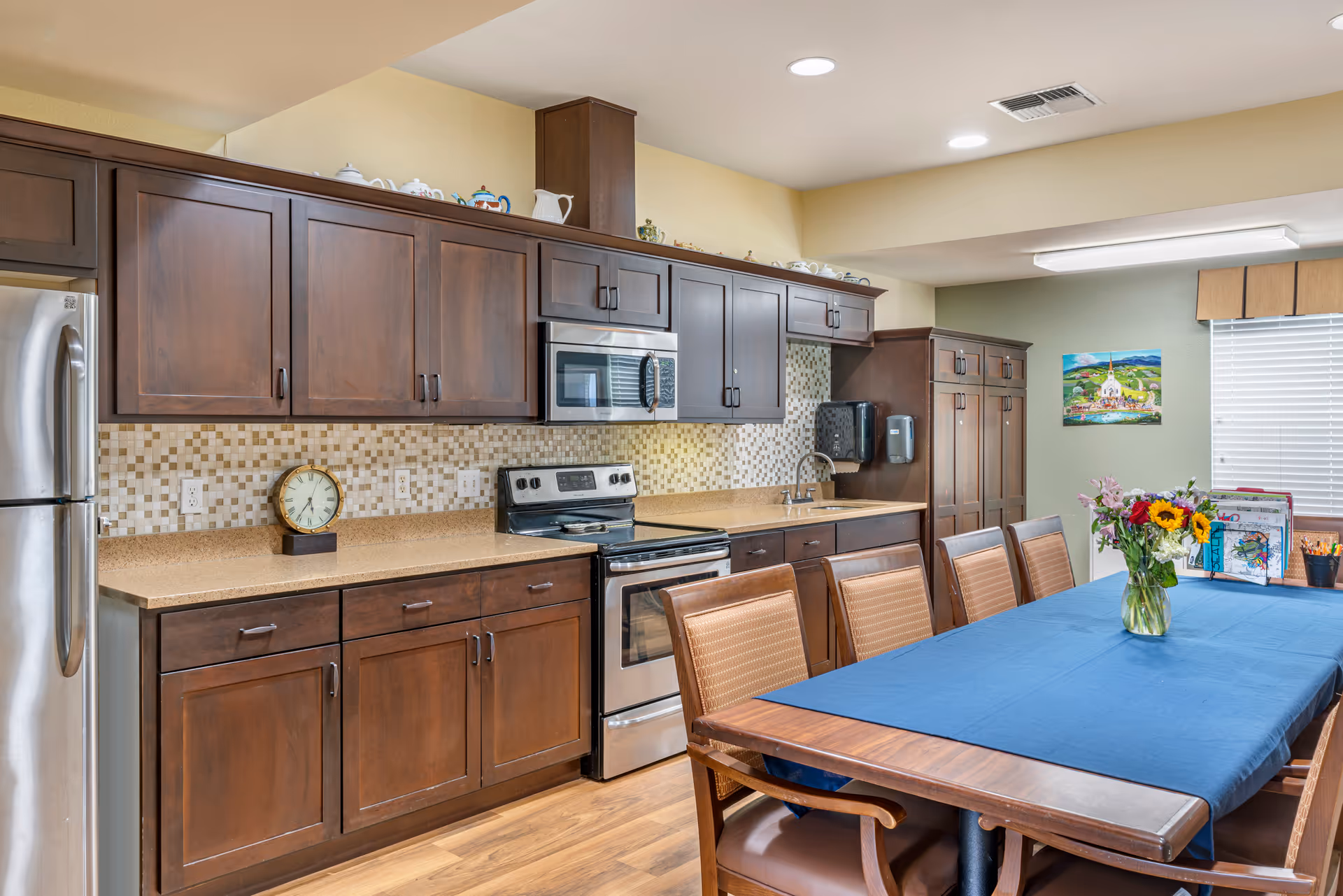 A kitchen and dining area with dark wooden cabinets, a stainless steel refrigerator, stove, and microwave. The backsplash features a mosaic tile design. A long wooden dining table with a blue table runner and six chairs is in the foreground, with a vase of flowers and some artwork on the table. The room has wood flooring and a window with blinds and a valance.