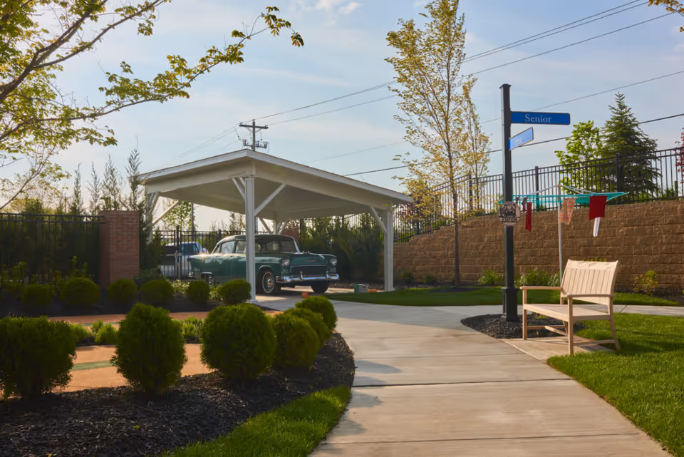 Outdoor area of a senior living facility with a paved walkway, neatly trimmed bushes, a wooden bench, a covered parking area with a vintage car, and a street sign labeled 'Senior'. Trees and a brick retaining wall are visible in the background under a clear sky.