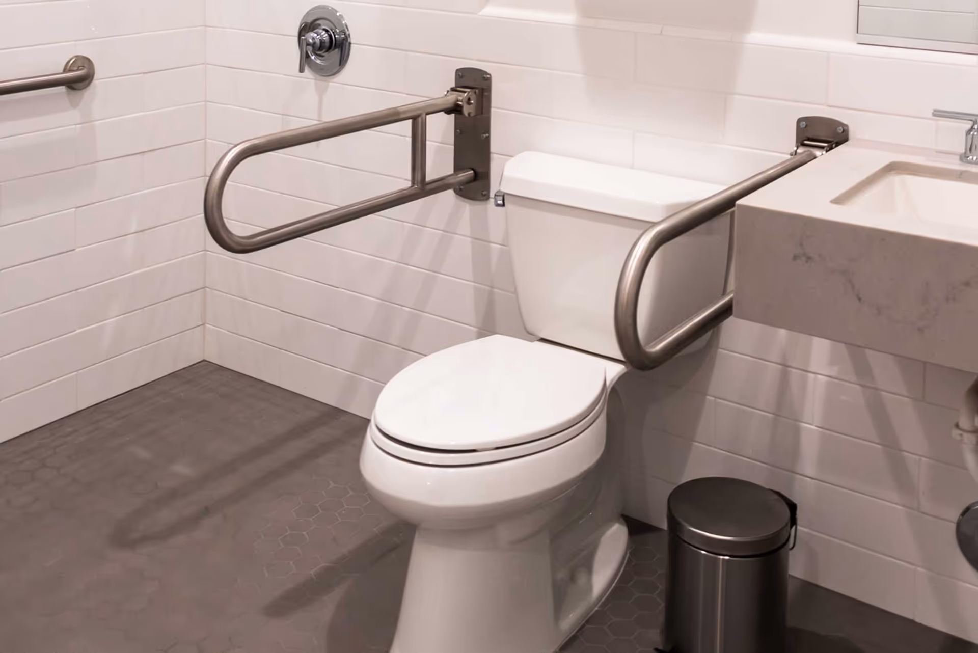 A clean, accessible bathroom featuring a white toilet with stainless steel grab bars on both sides, white tiled walls, a gray hexagonal tiled floor, a white sink with a marble countertop, and a small stainless steel trash can.