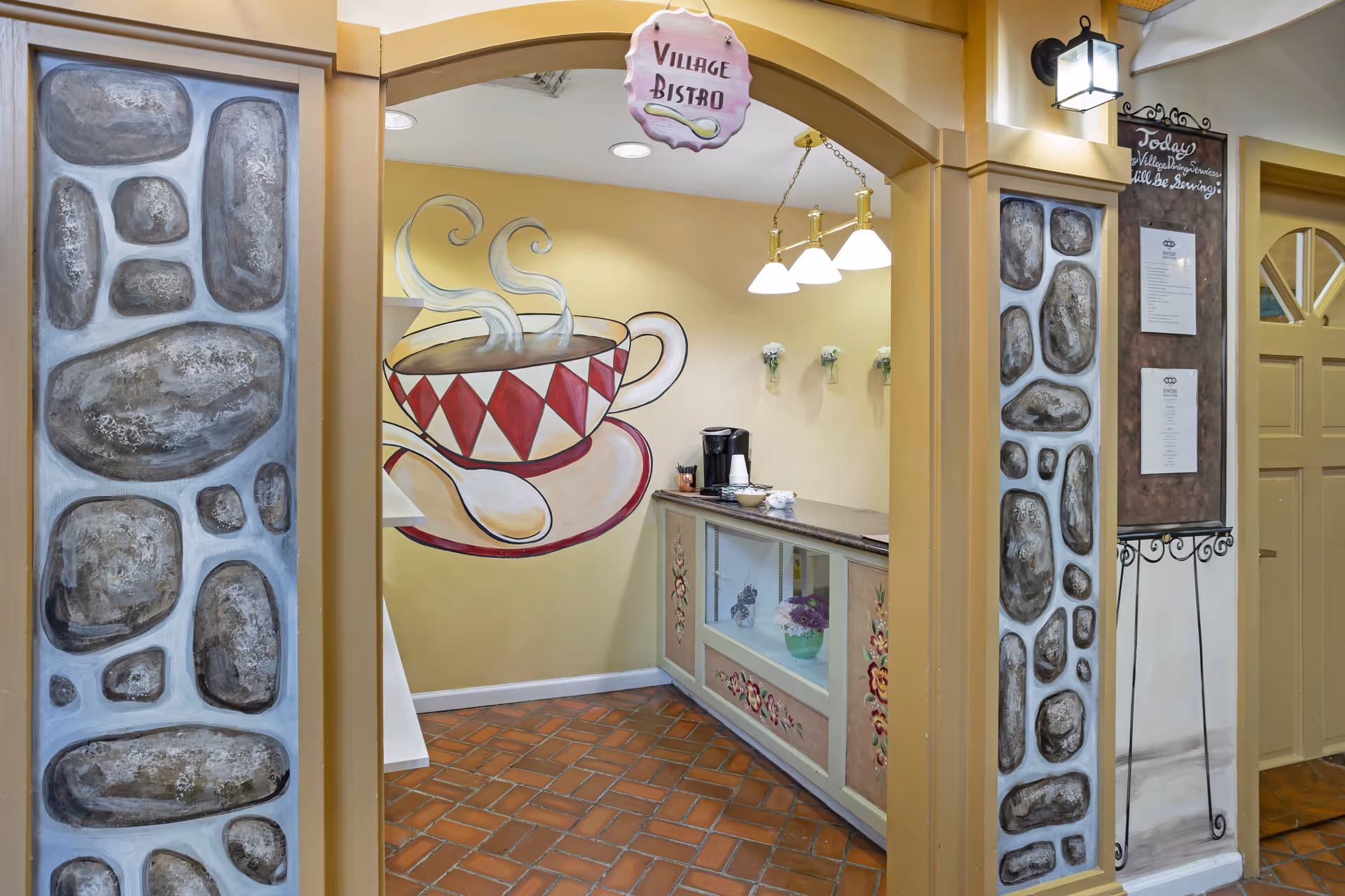Interior view of a small bistro area named Village Bistro with a painted mural of a steaming cup of coffee on the wall. The space features a counter with a coffee machine, cups, and utensils. The walls have decorative stone-like paintings and a hanging sign that reads 'Village Bistro'. There is also a chalkboard menu on a stand to the right.