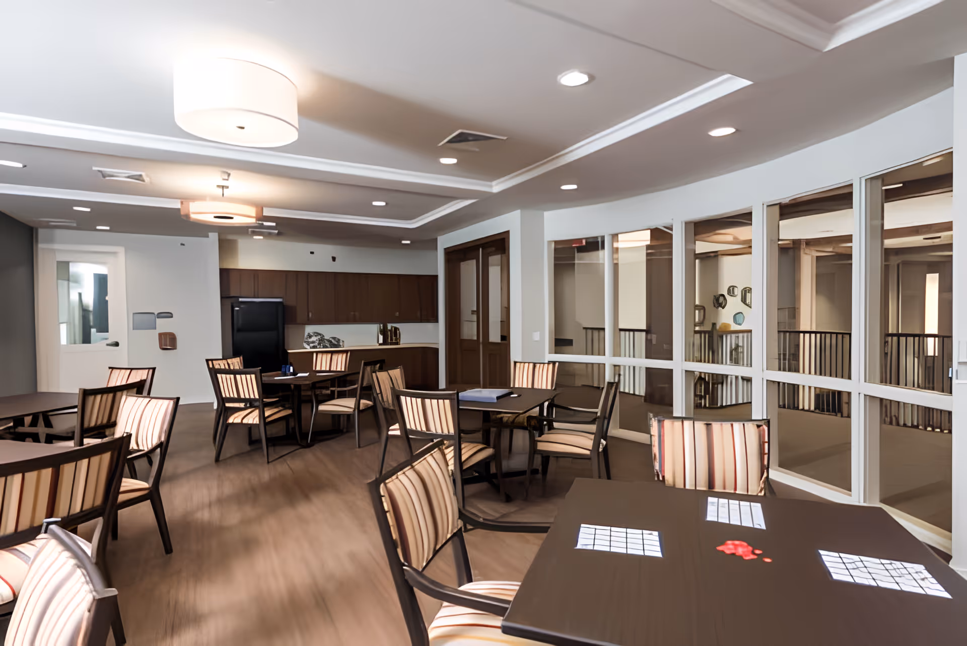Communal dining/activity room with multiple tables and striped chairs, a kitchenette area, and large interior windows.